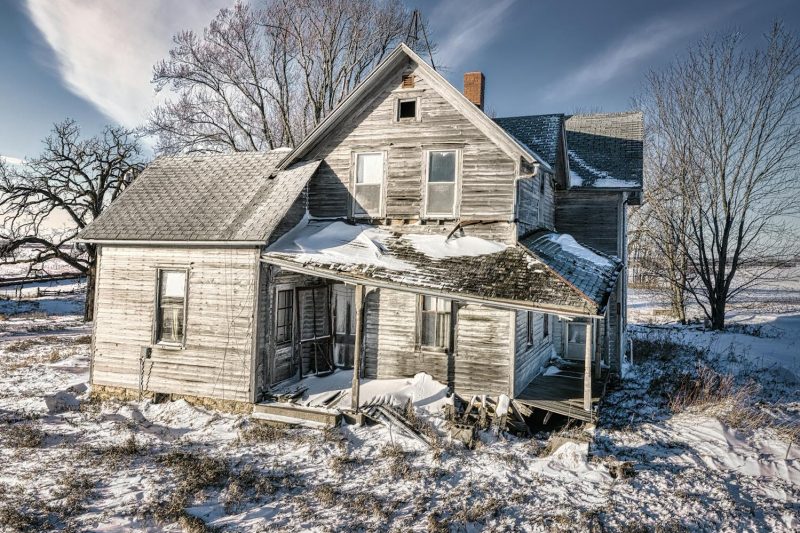 Abandoned wooden house covered in snow, surrounded by bare trees under a clear blue sky.