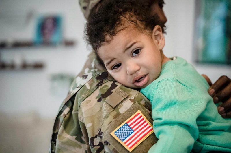 Child in a green sweater hugging military parent with a U.S. flag patch, showcasing a heartwarming bond.