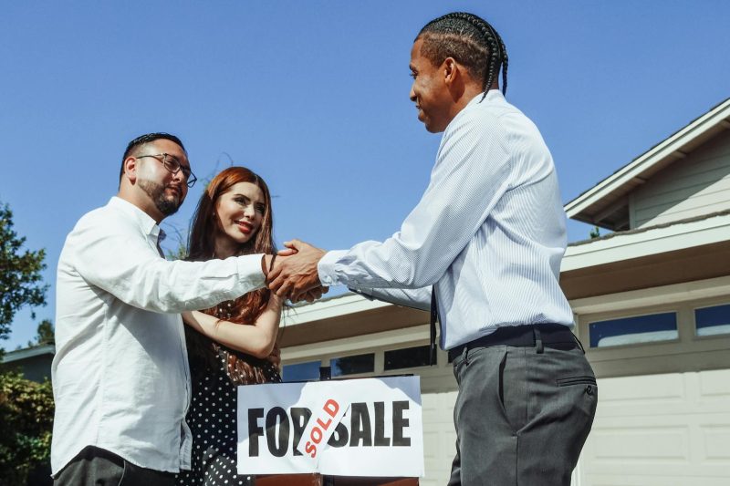 A couple shakes hands with their realtor in front of a newly purchased home, smiling and joyful.