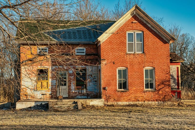 Abandoned rustic brick house with peeling paint, surrounded by bare trees and fallen leaves during late fall.