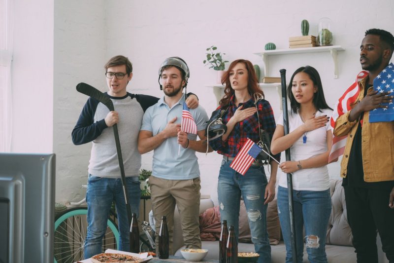 Diverse friends gathered in a cozy living room, watching sports on TV with American flags displayed indoors.