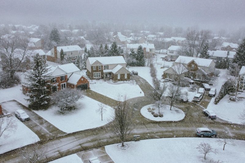 Aerial view of a snowy suburban neighborhood featuring houses, trees, and quiet streets under a serene winter sky.