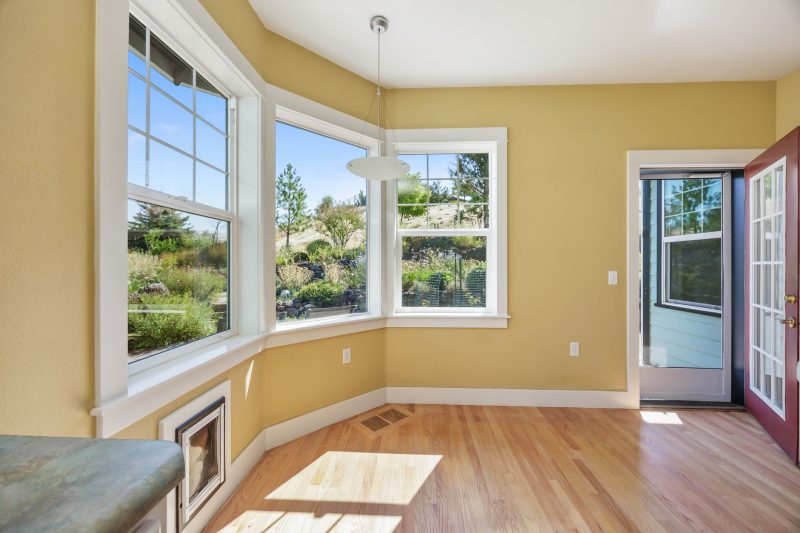 Spacious sunlit room with wooden floors and large windows offering views of a garden.
