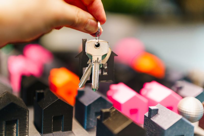 Hand holding keys above colorful miniature houses, symbolizing real estate choices.
